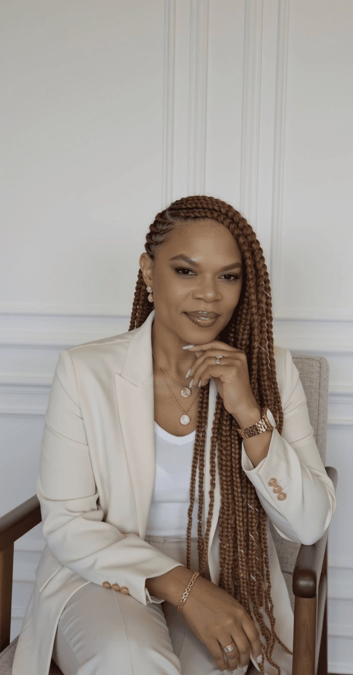 Smiling woman with long brown braids wearing a cream blazer and gold jewelry, seated.