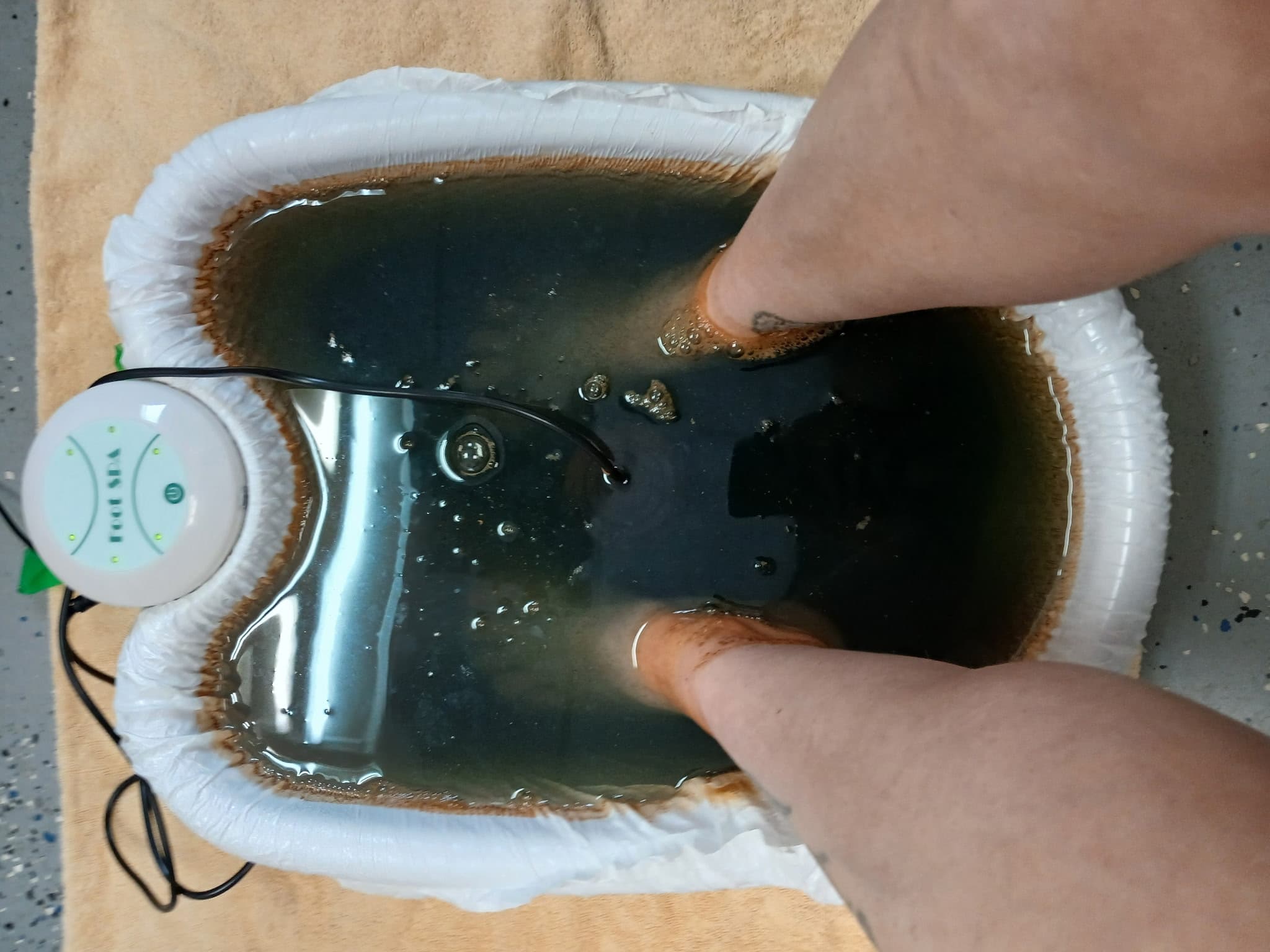 A person's feet soaking in dark, murky water within an ionic detox foot bath basin.