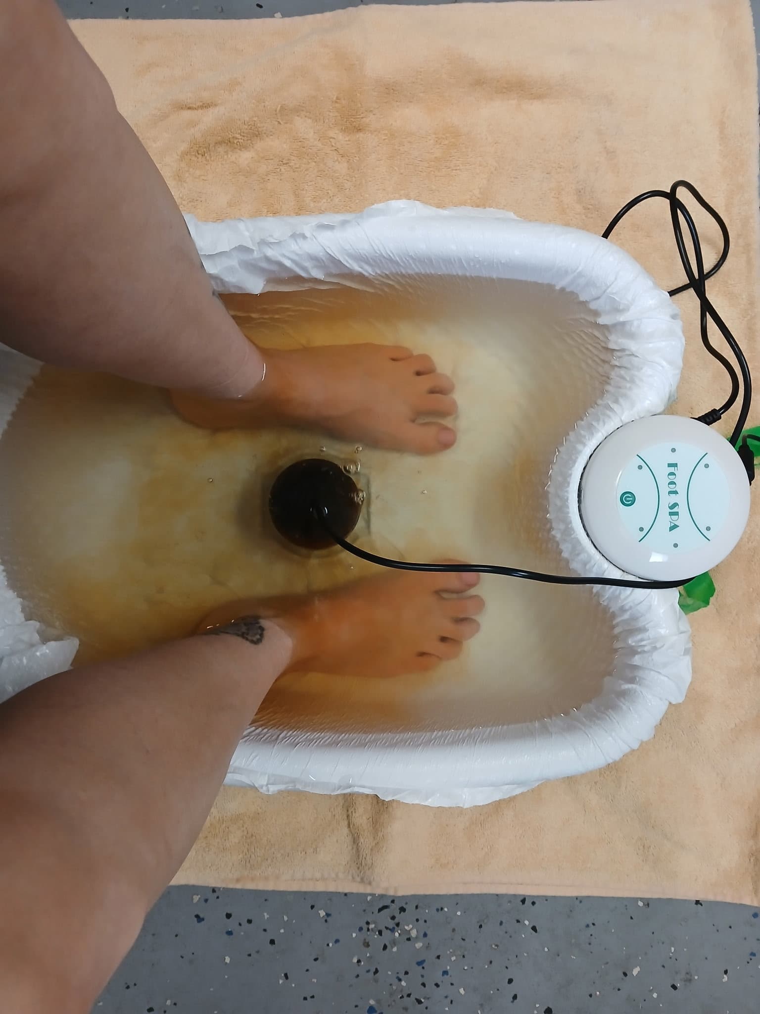 Feet soaking in a white ionic detox foot spa basin filled with murky brown water.